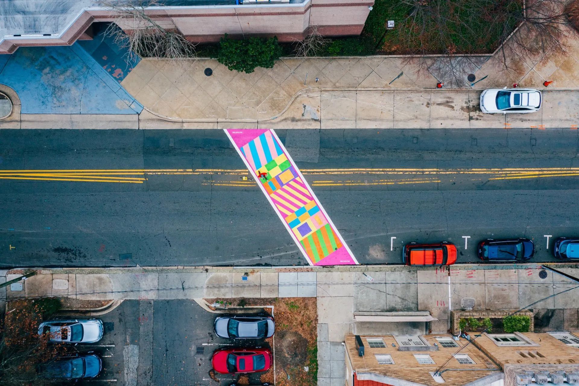 An aerial view of a vibrant, multi-colored, geometric-patterned pedestrian crosswalk painted on a grey asphalt city street.