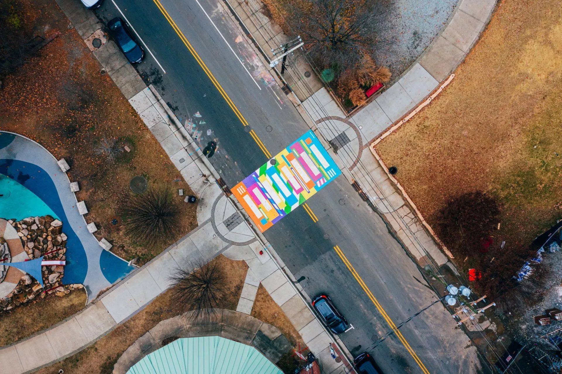 An aerial view of a vibrant, multicolored crosswalk painted on an asphalt street between sidewalks and a small park.