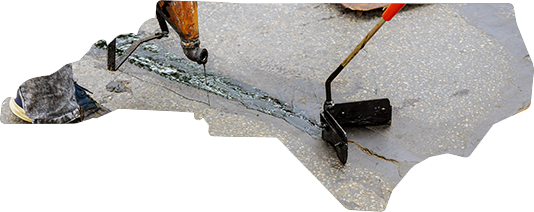 A worker uses a handheld tool to apply black sealant into a crack on a grey pavement surface.