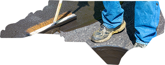 A person in blue jeans uses a push broom to spread a dark liquid sealant over an asphalt surface shaped like North Carolina.