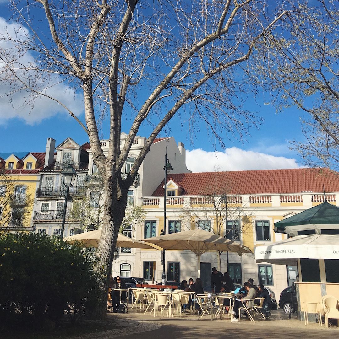 A group of people are sitting under umbrellas in front of a building. Principe Real Square, Lisbon.