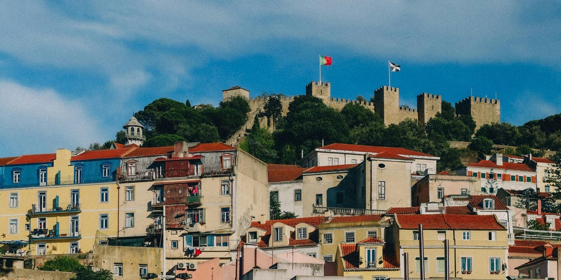 A majestic view of São Jorge Castle in Lisbon, perched atop a hill with panoramic vistas of the city below. The medieval fortress, with its sturdy stone walls, towers, and watchtowers, stands as a symbol of Lisbon’s rich history. Surrounded by lush gardens and offering sweeping views of the Tagus River and iconic landmarks, the castle provides a glimpse into Lisbon's past and breathtaking cityscape.
