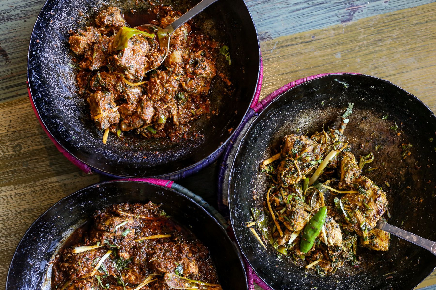 Three bowls of food with spoons in them are on a wooden table.