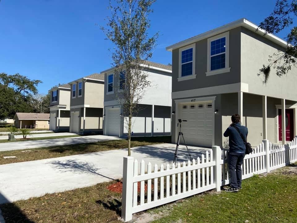 A man is standing in front of a white picket fence in front of a row of houses.