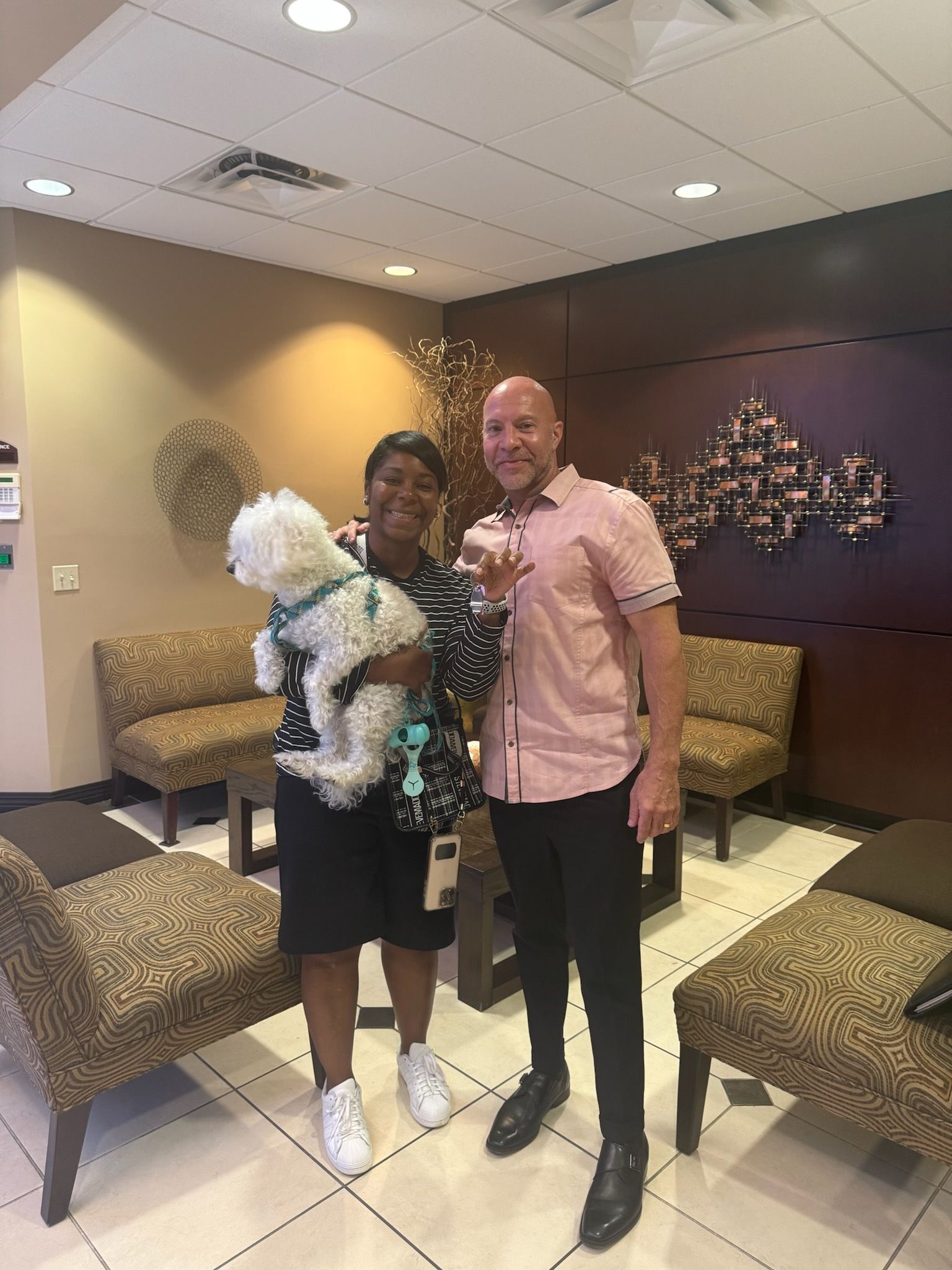 Woman, man, and dog in a waiting room. Woman holds fluffy white dog. Man smiles, hand raised. Neutral-toned setting.