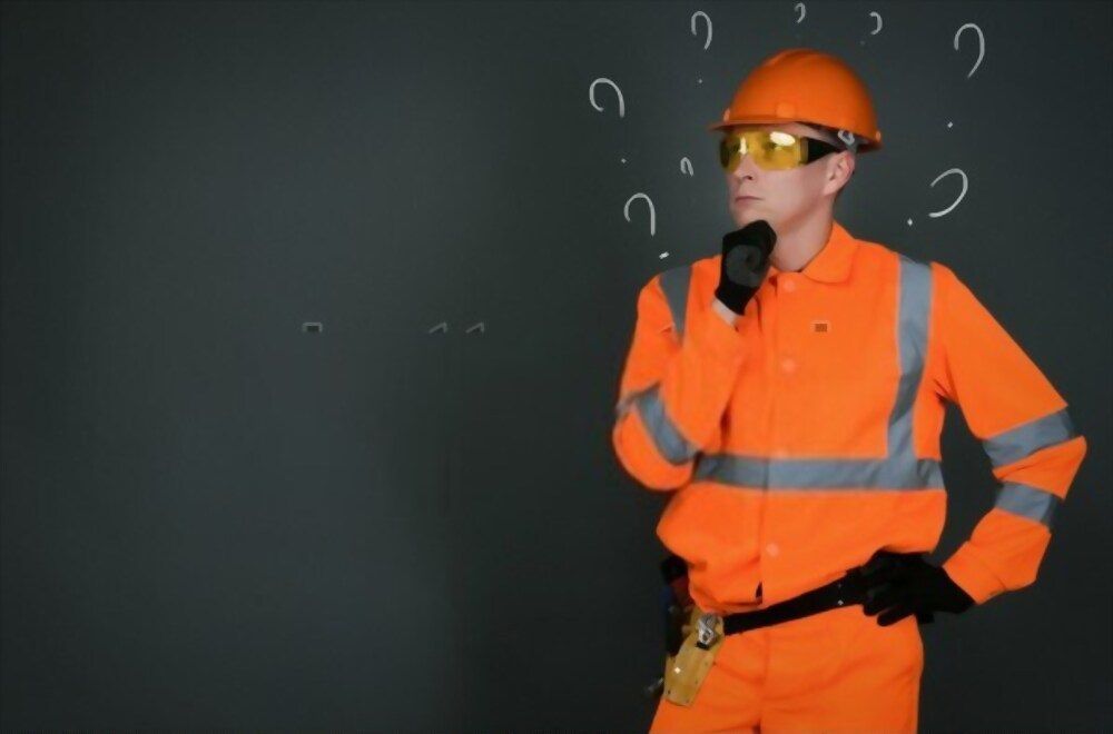 A construction worker is standing in front of a blackboard with question marks written on it.