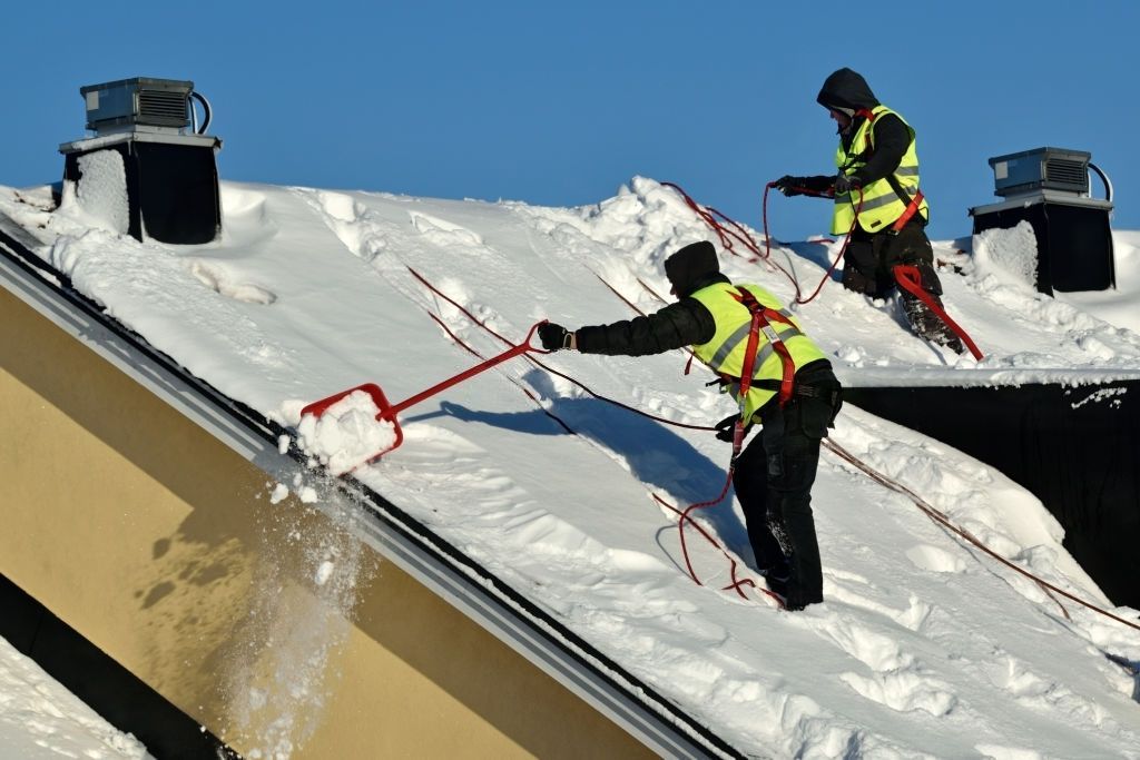 Two men are clearing snow from the roof of a building