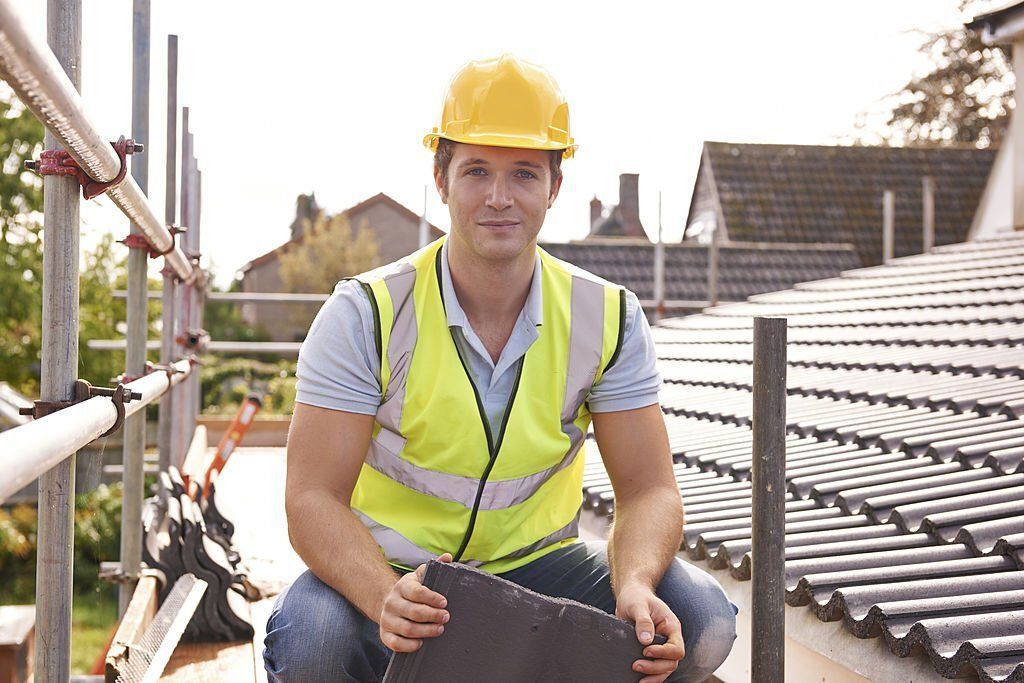 A construction worker is sitting on a roof holding a piece of slate.