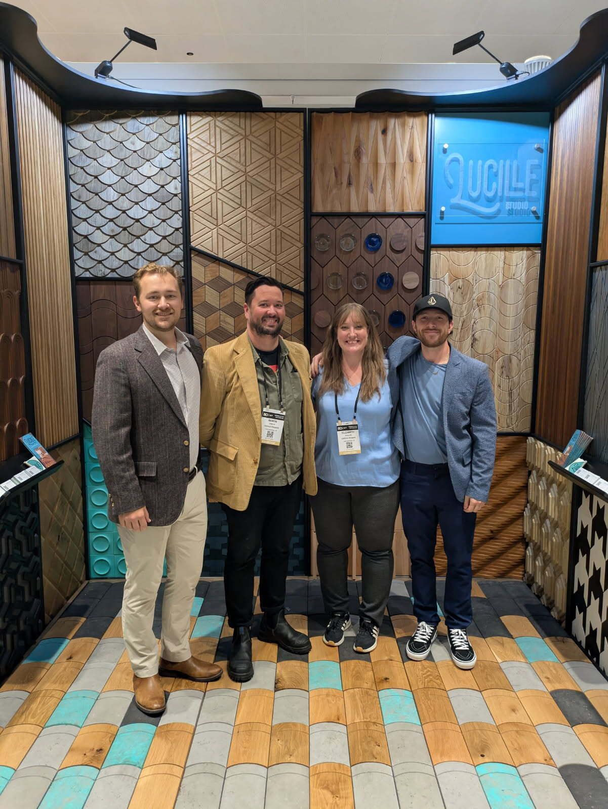 Four people pose in front of a patterned wall display. Various textured wood panels and a tile floor.