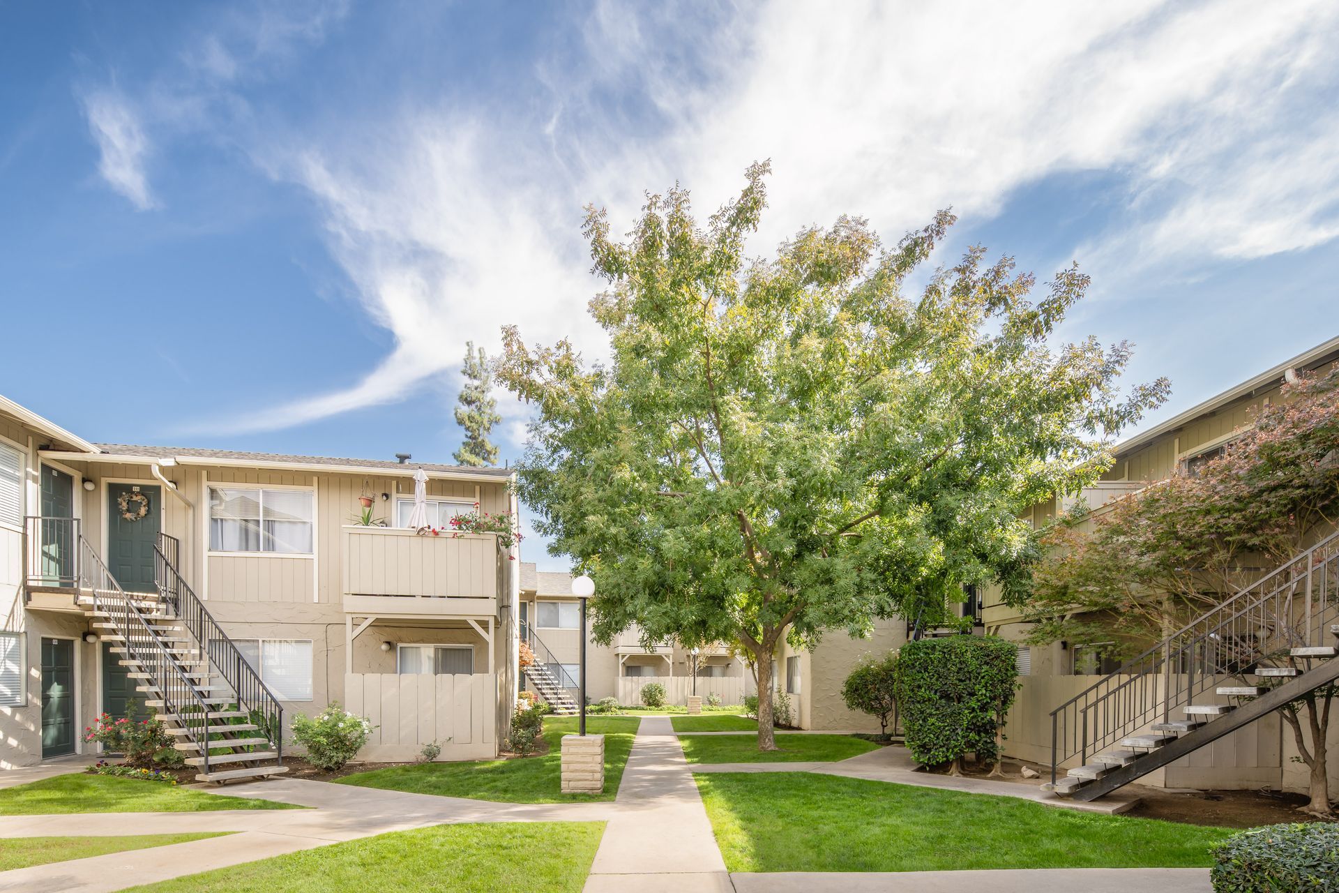 Two-story apartment complex with a pathway and central tree under a blue sky.