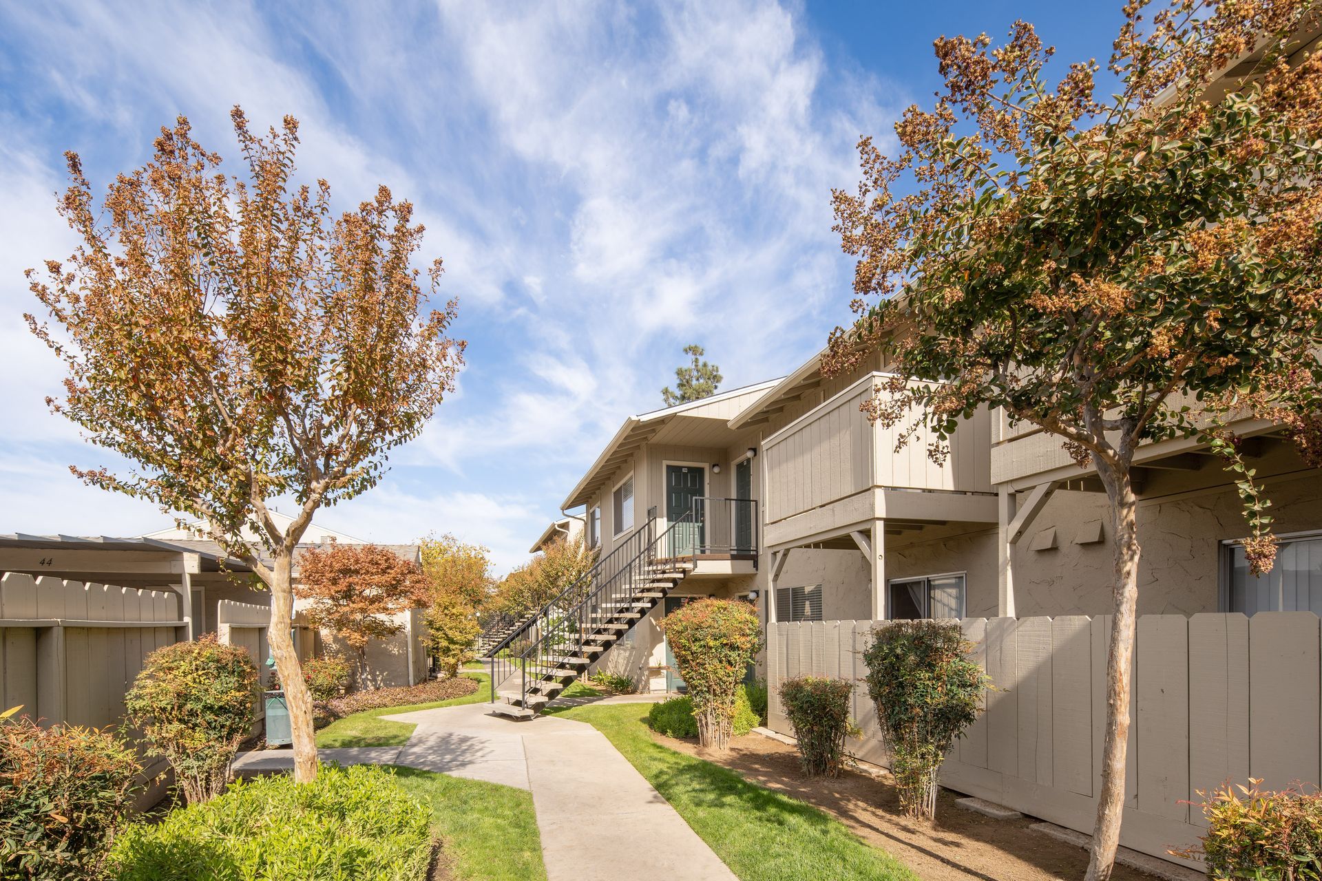 Apartment complex exterior with trees and a walkway under a blue sky.