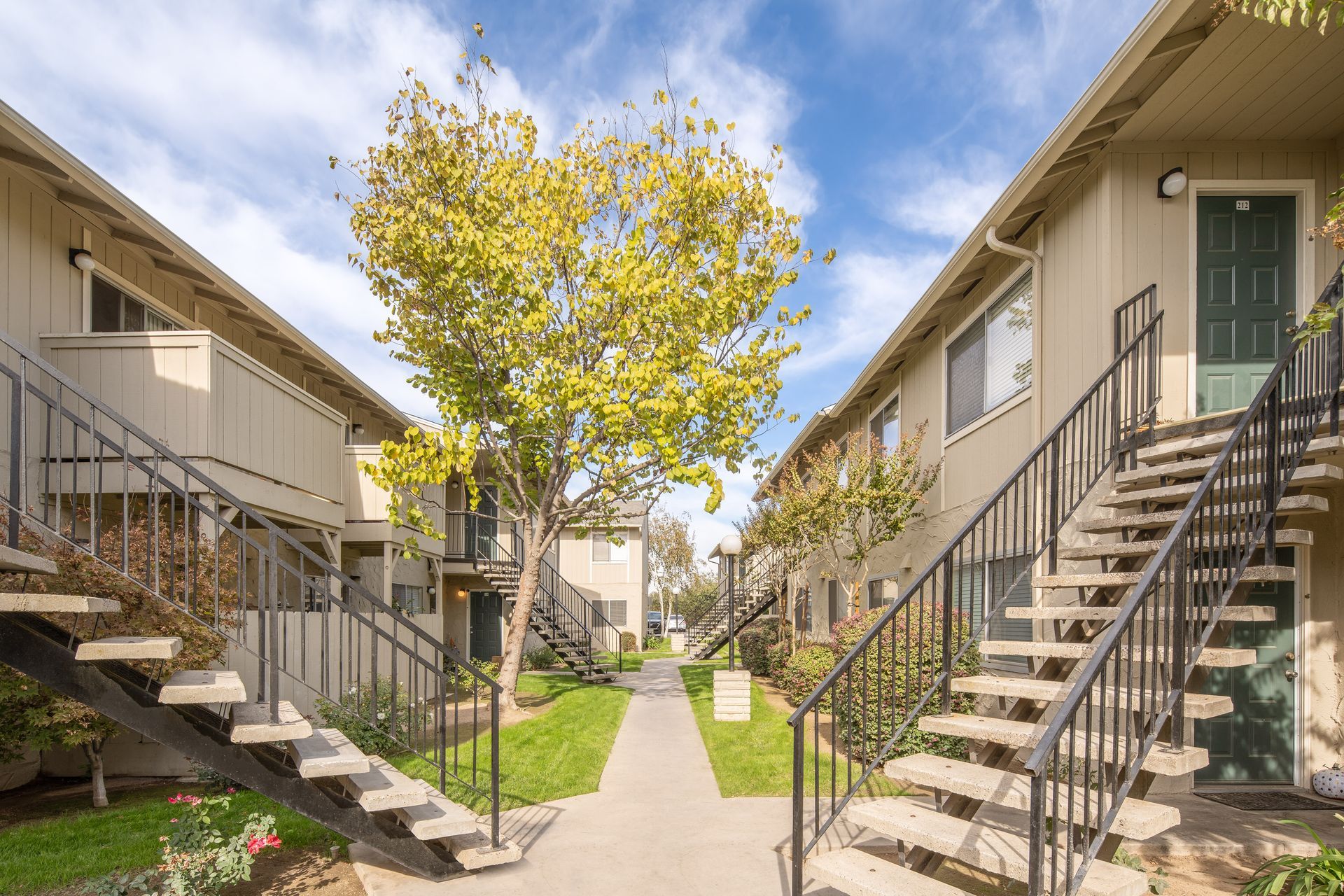 Apartment complex with exterior staircases, central walkway, tree, and blue sky.