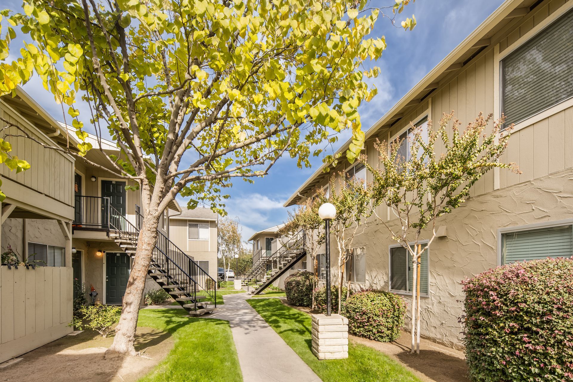 Apartment complex with beige buildings, green grass, trees, and a blue sky.