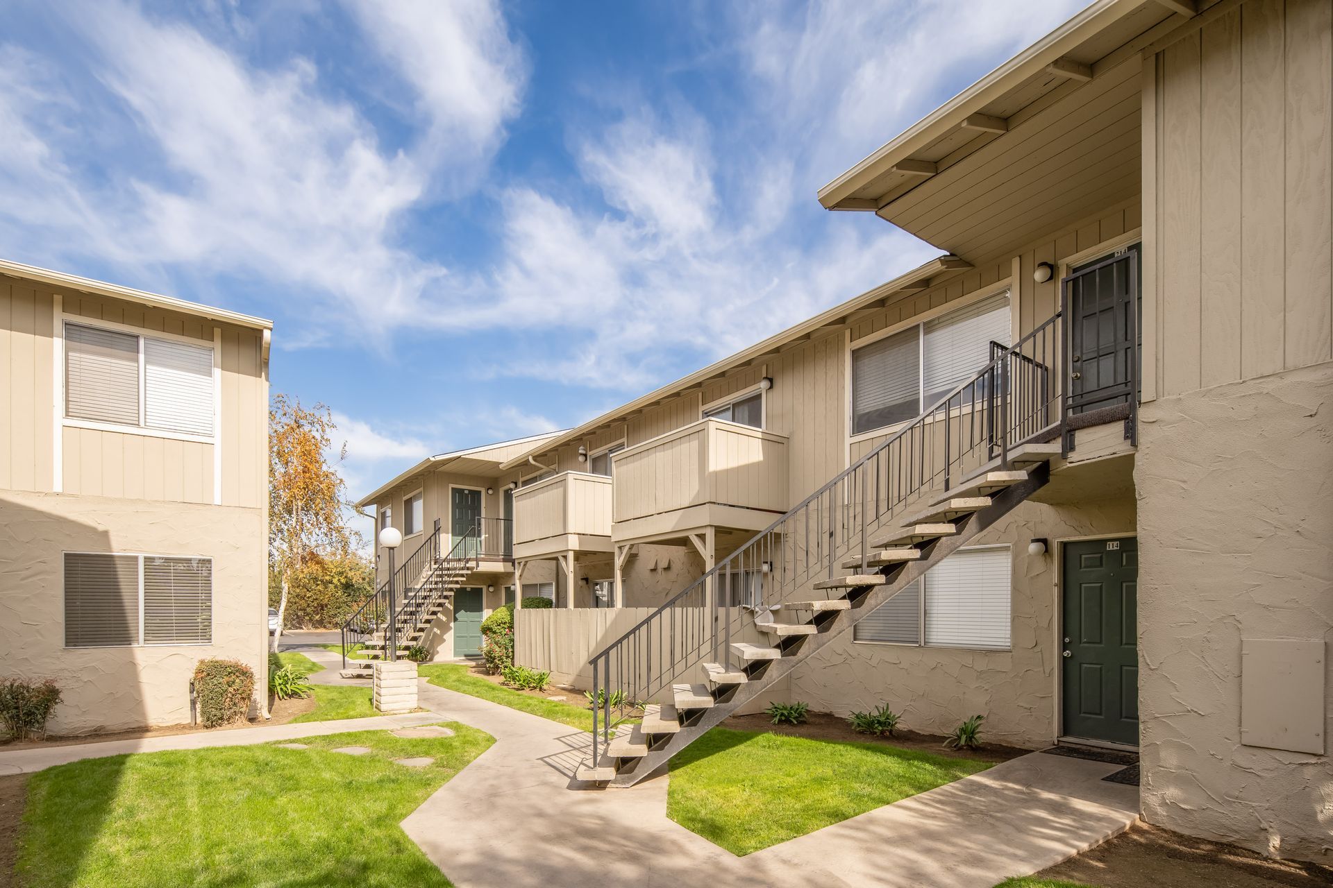 Apartment complex with beige buildings, stairs, green grass, and a blue sky with clouds.