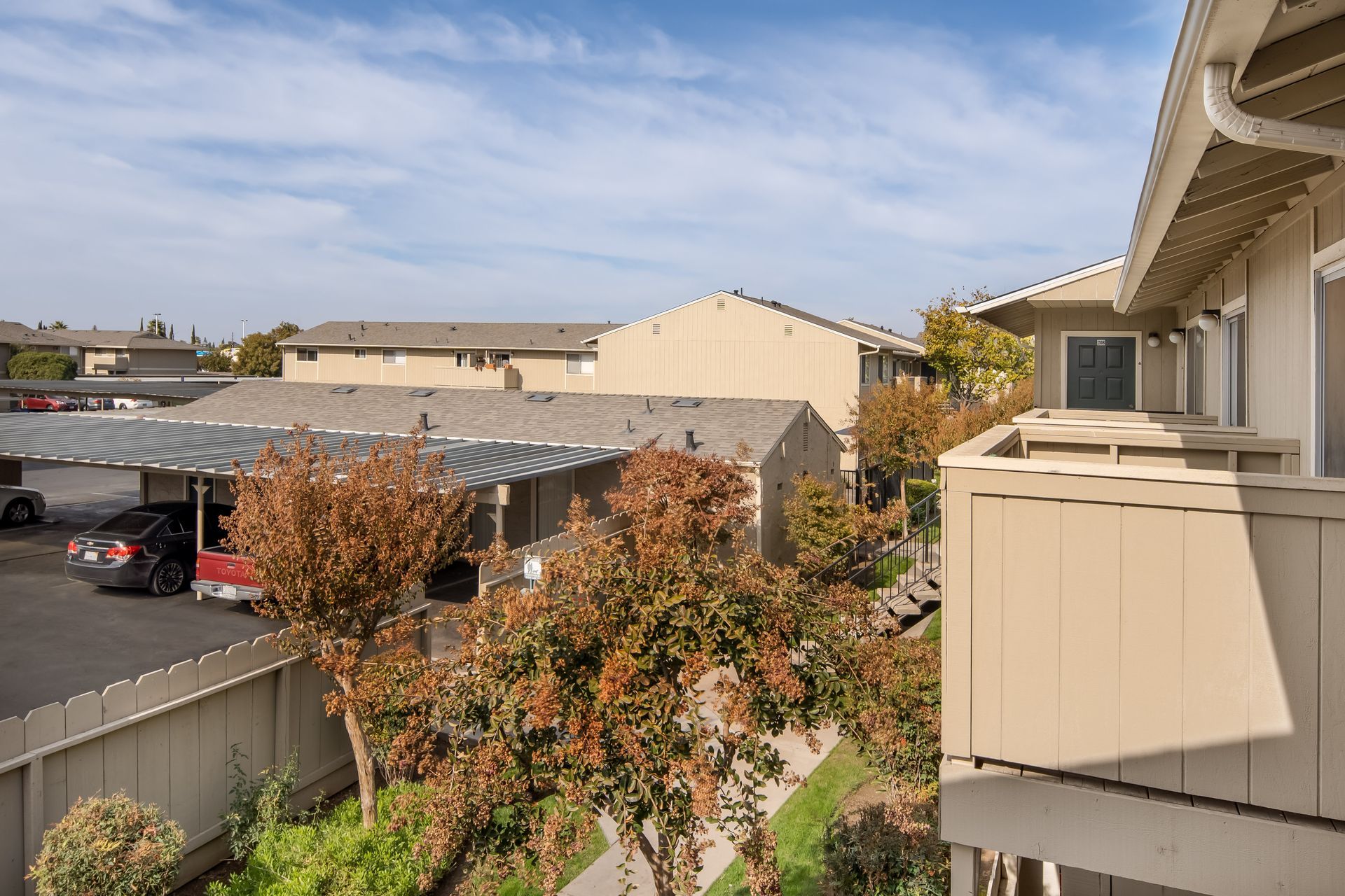 Apartment complex with tan buildings, covered parking, and trees under a blue sky.