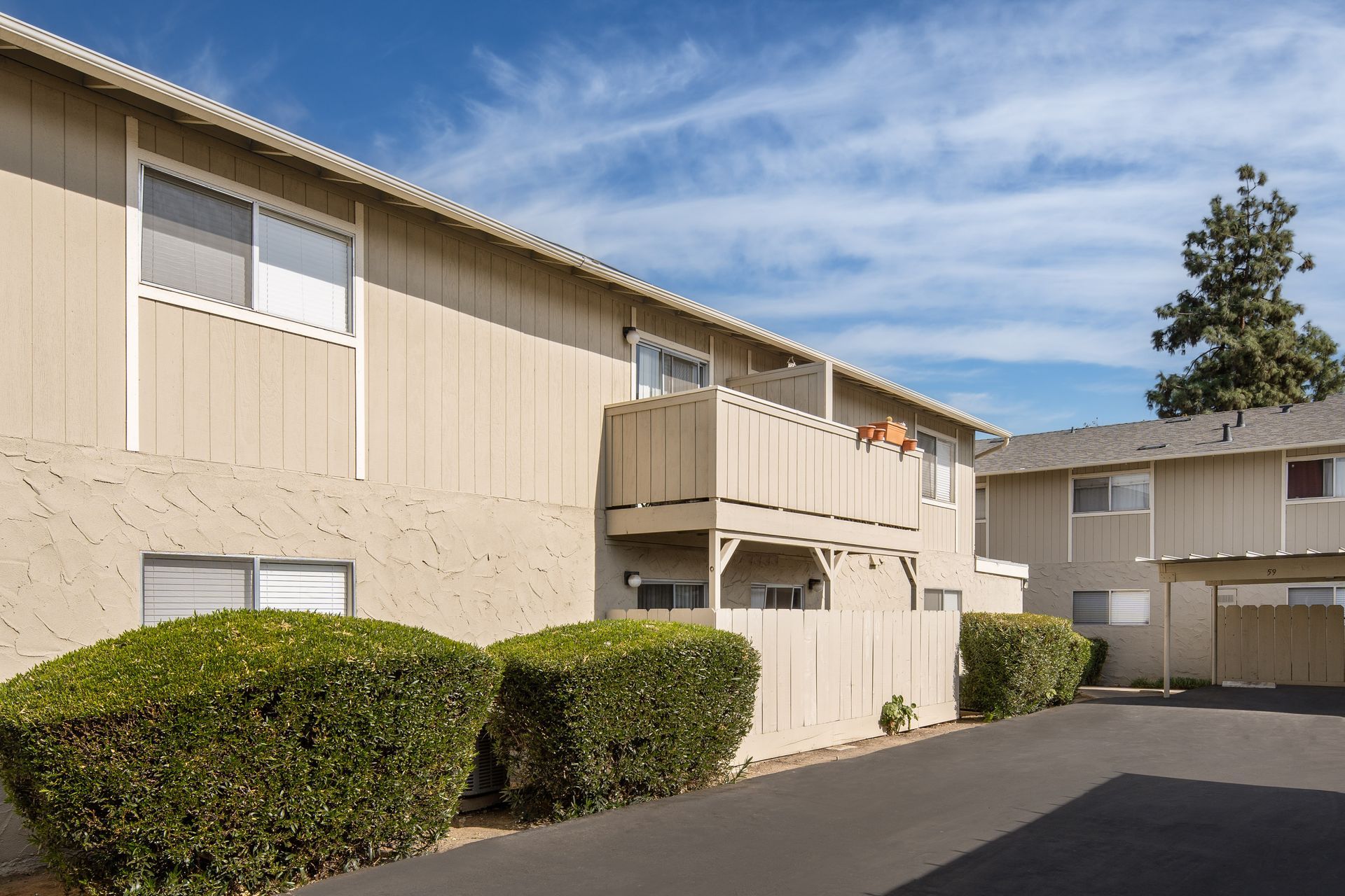 Two-story beige apartment complex with a balcony, bushes, and blue sky.