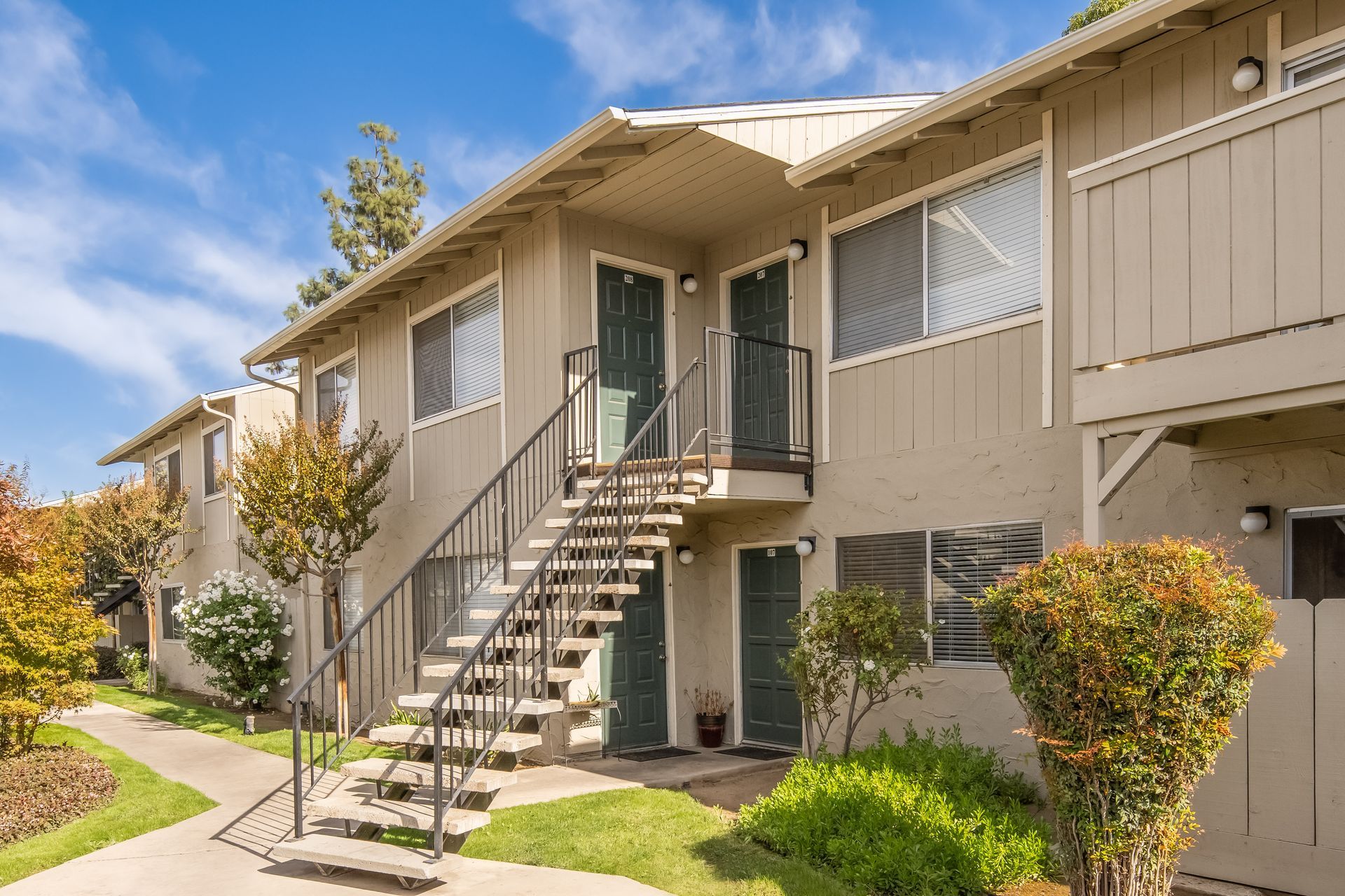 Two-story apartment building with stairs leading to upper units; beige exterior, green doors, blue sky.