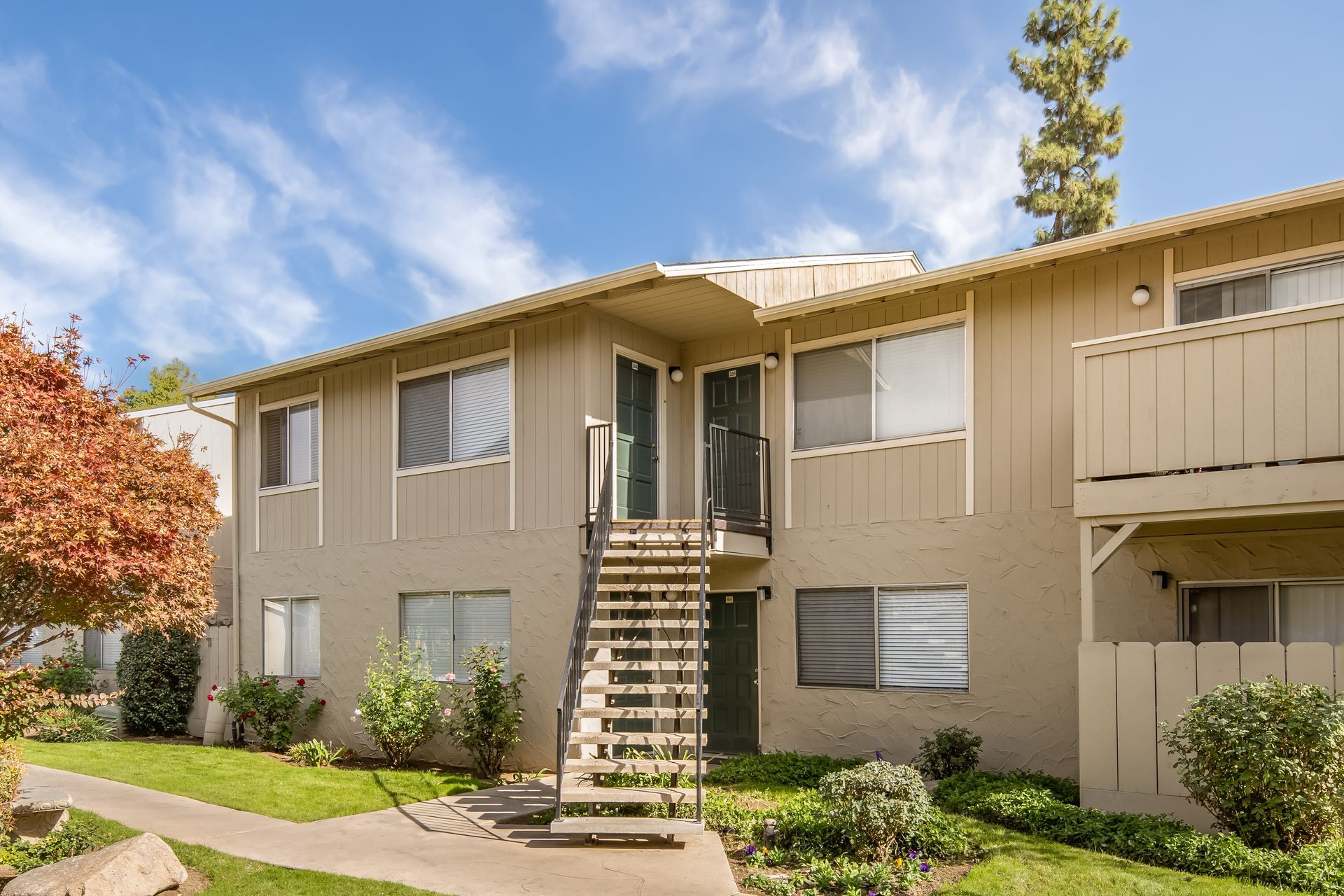 Two-story beige apartment building with exterior stairs, greenery, and blue sky.