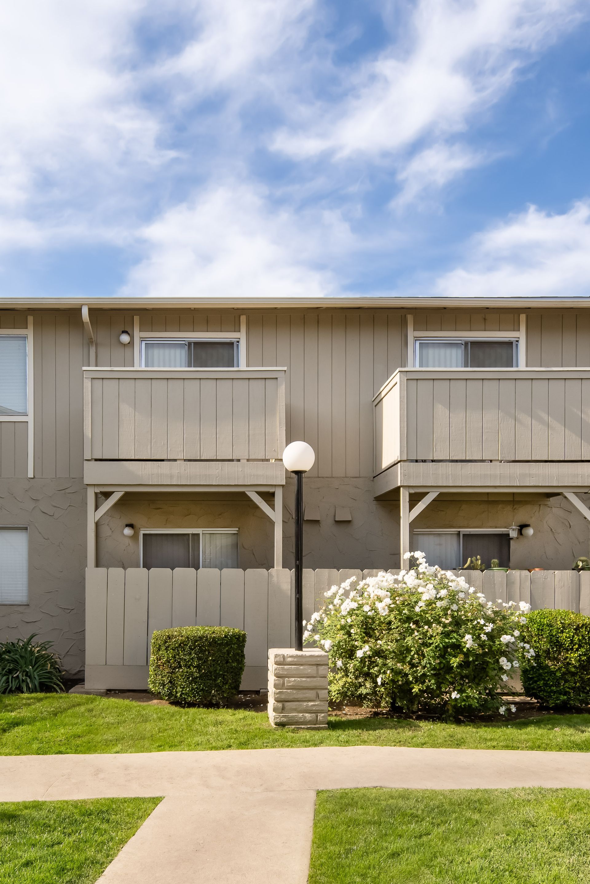 Two-story tan apartment building with balconies, a sidewalk, green lawn, and a sunny sky.