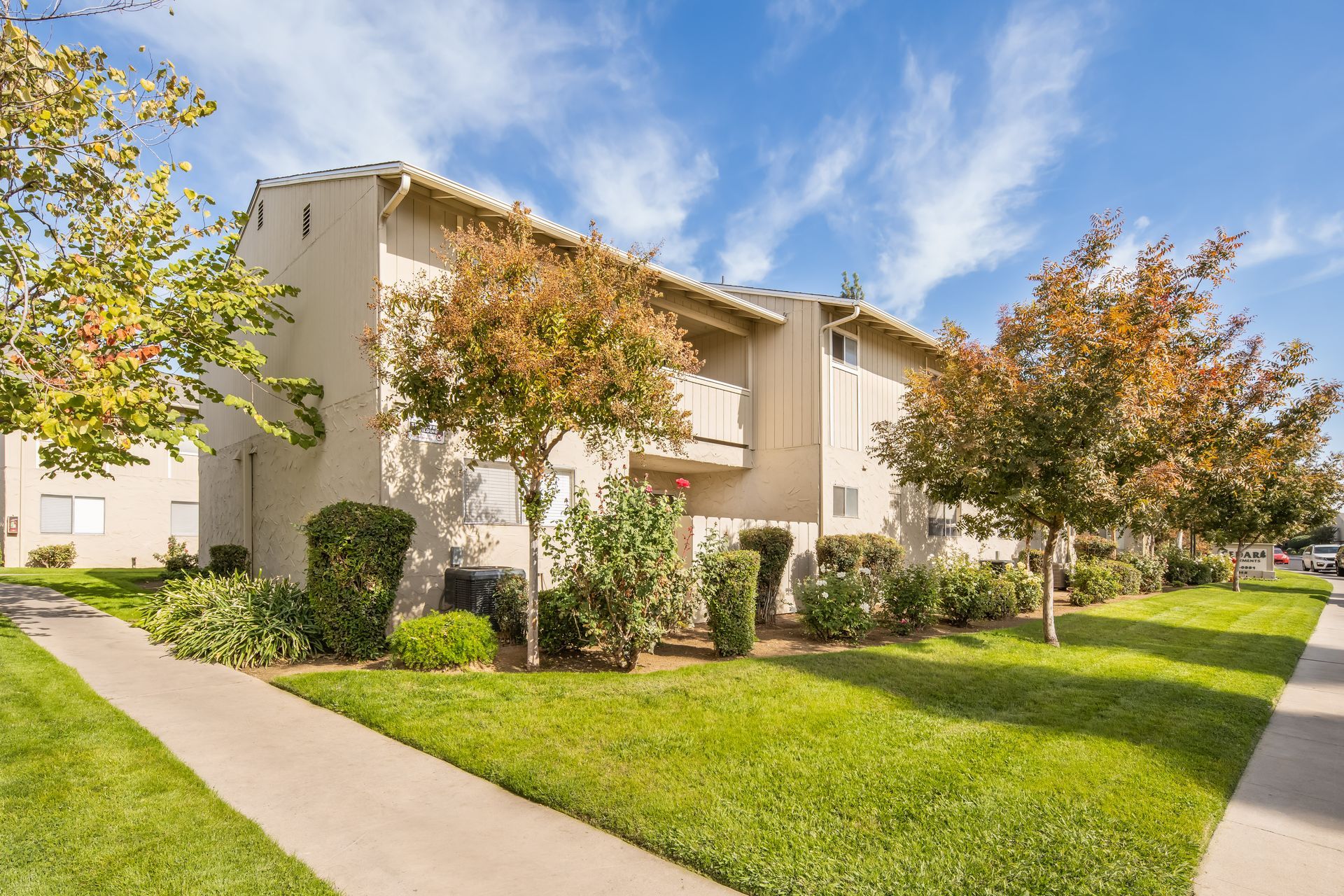 Apartment building with beige exterior, balconies, and green lawn with trees and sidewalk.