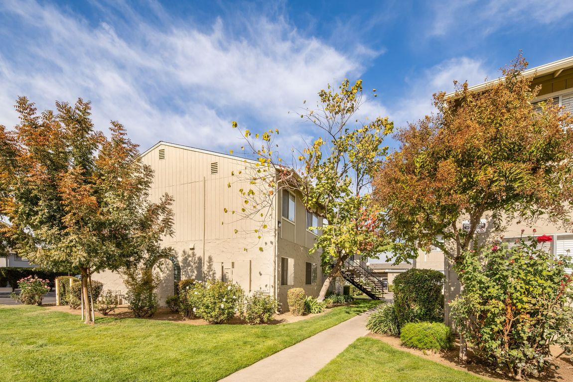 Apartment building with beige siding surrounded by trees and green grass under a blue sky.