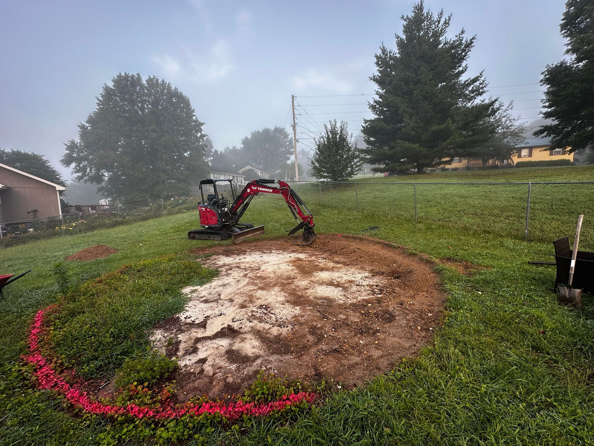 Mini excavator digging a circular area in a grassy yard on a foggy day. A house and trees are in the background.