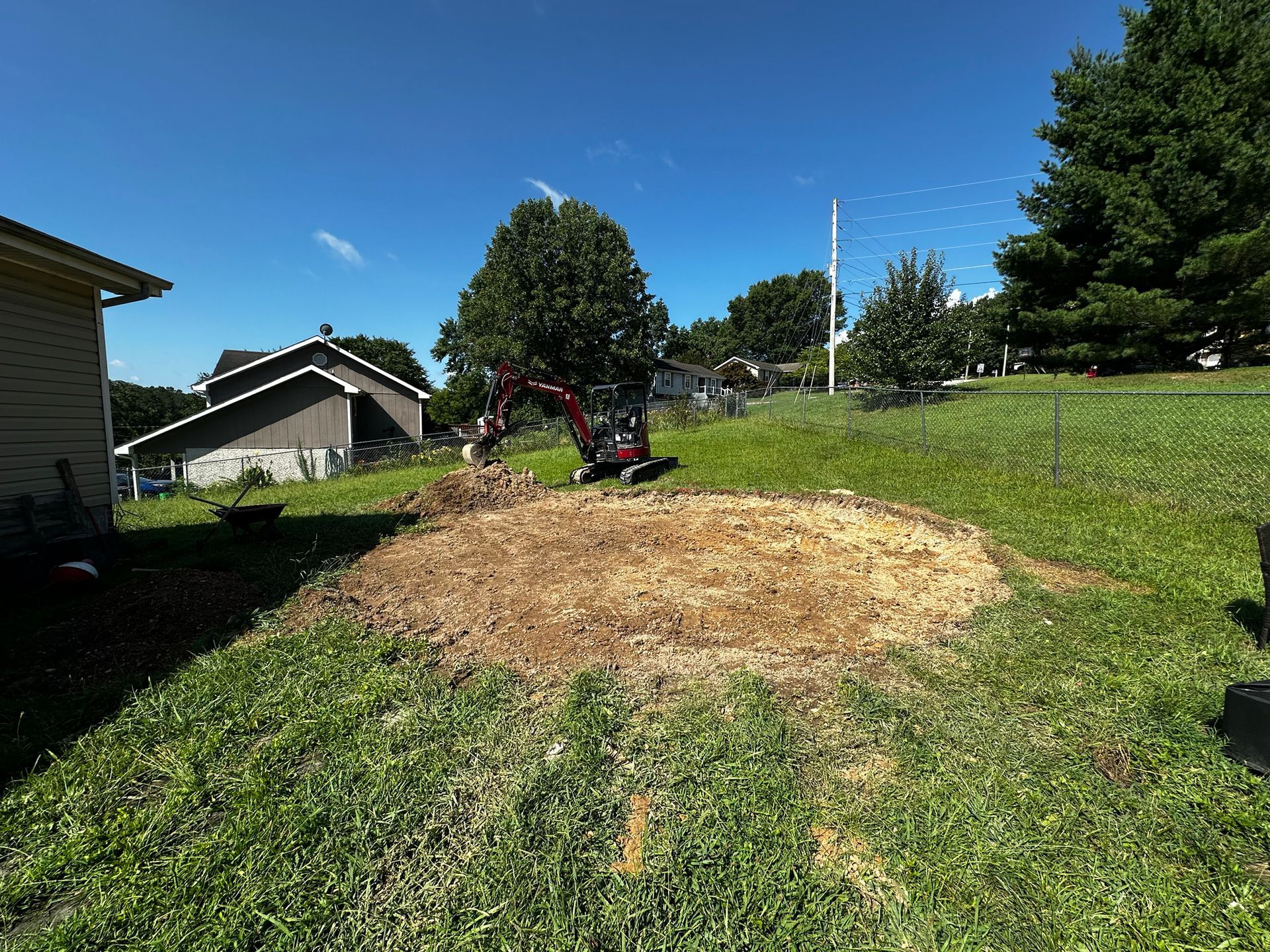 An excavator working on a patch of dirt in a grassy yard, under a blue sky.