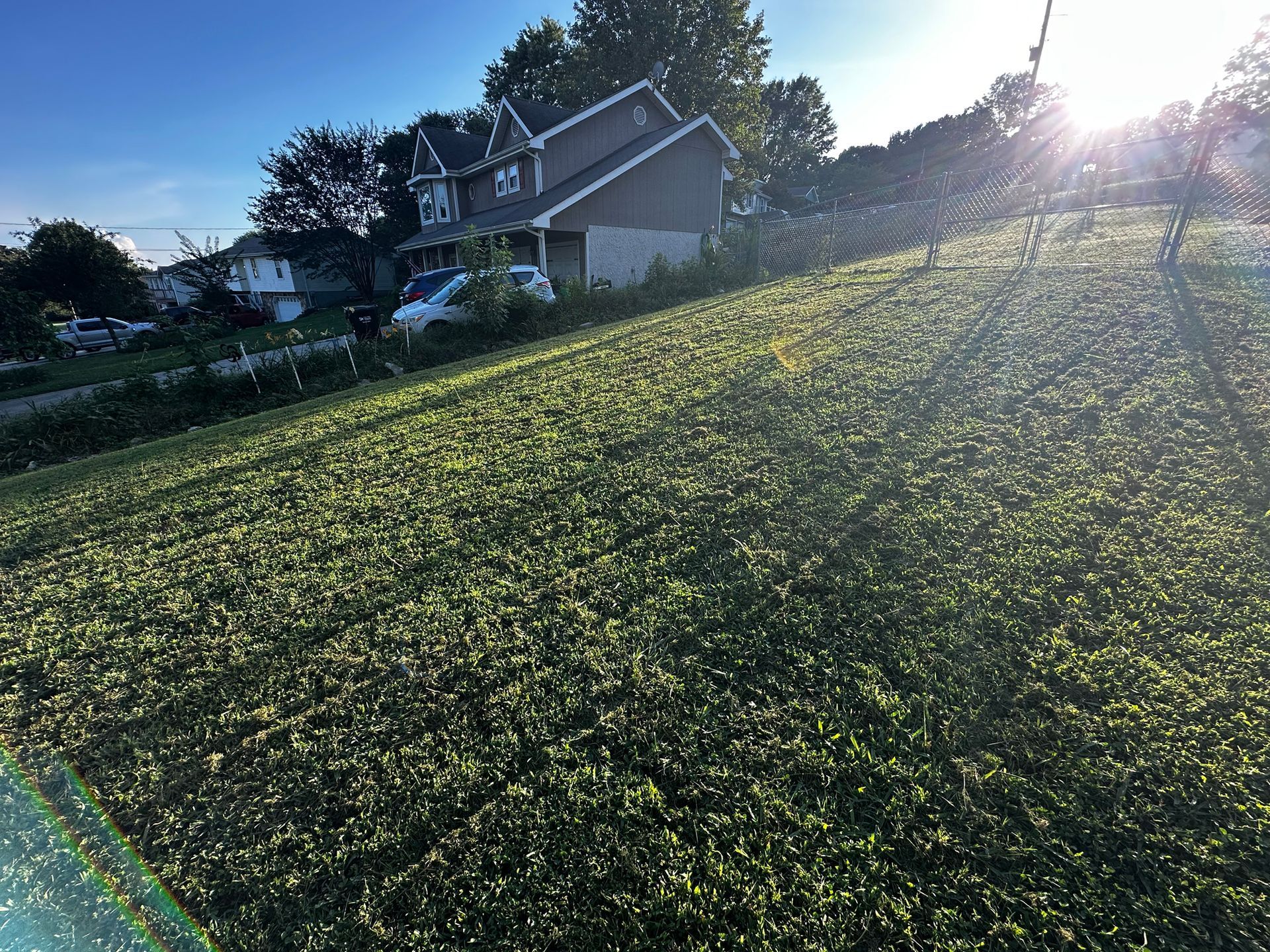 A sunlit grassy hill leads to a two-story house with a driveway and car. Blue sky in the background.