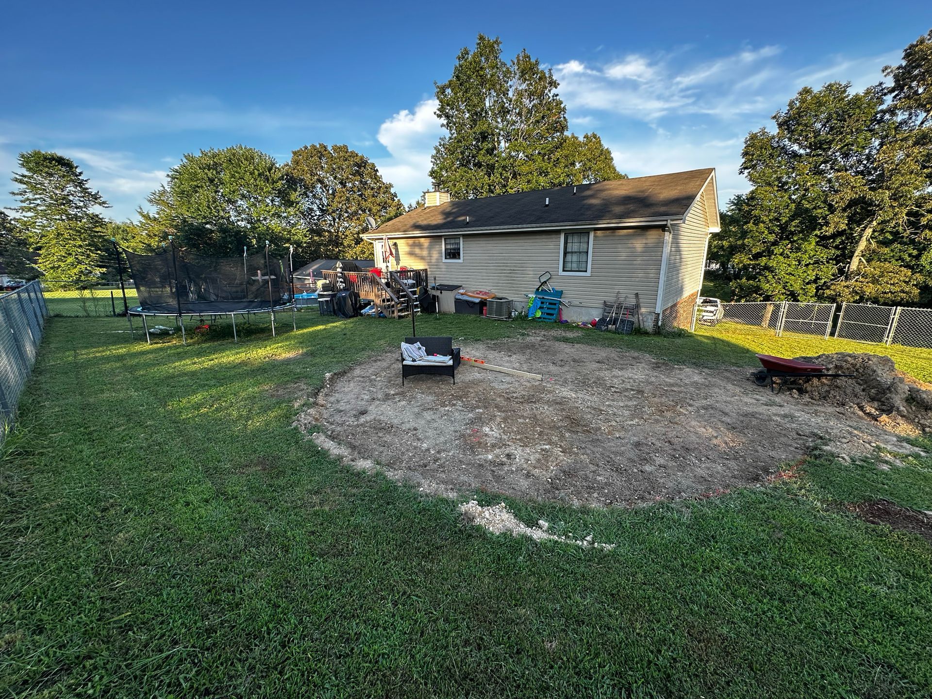 Backyard with a gravel area, grass, house, trampoline, and trees. Blue sky.
