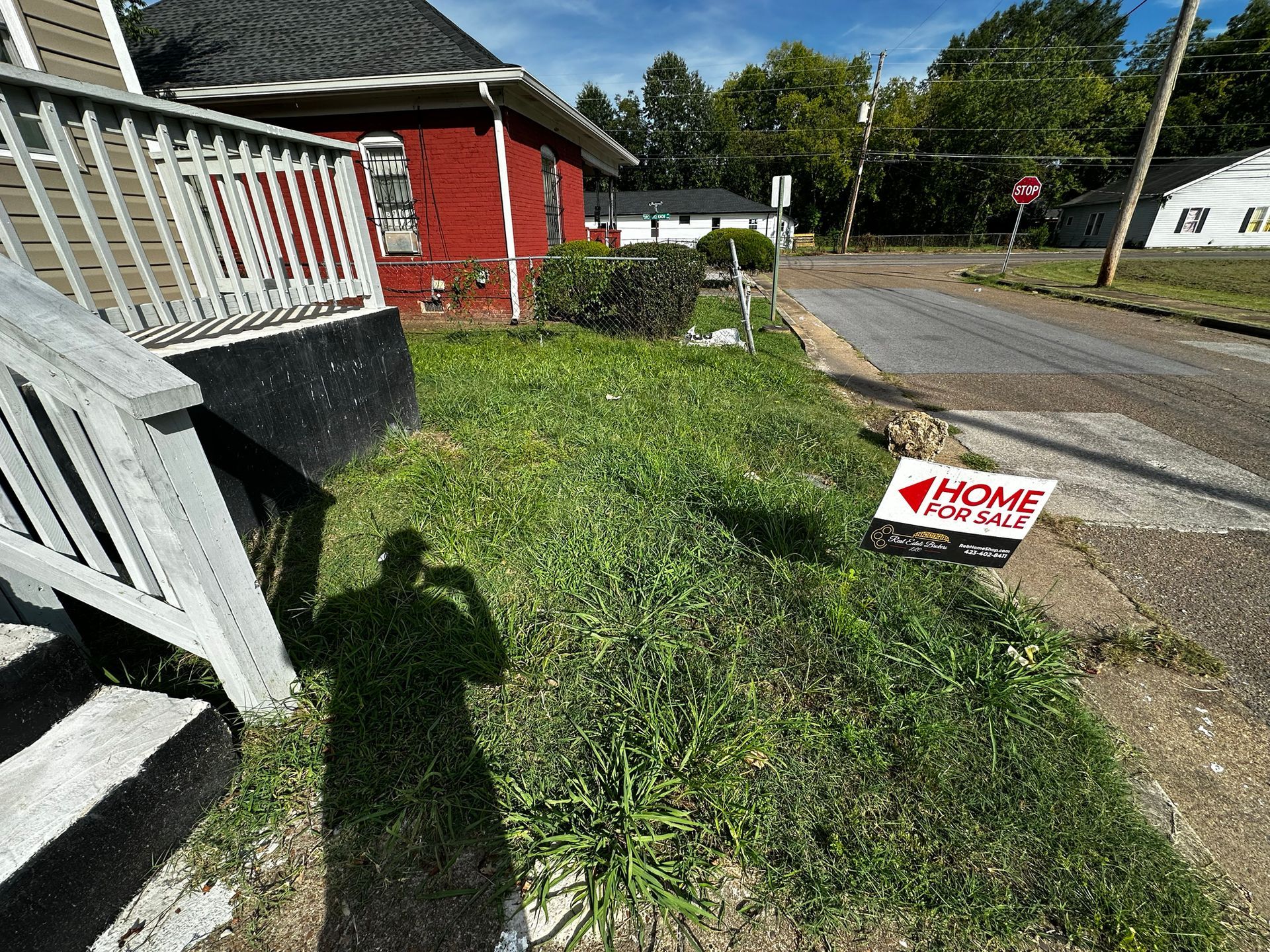 Home for sale sign on overgrown lawn next to a red house and sidewalk.
