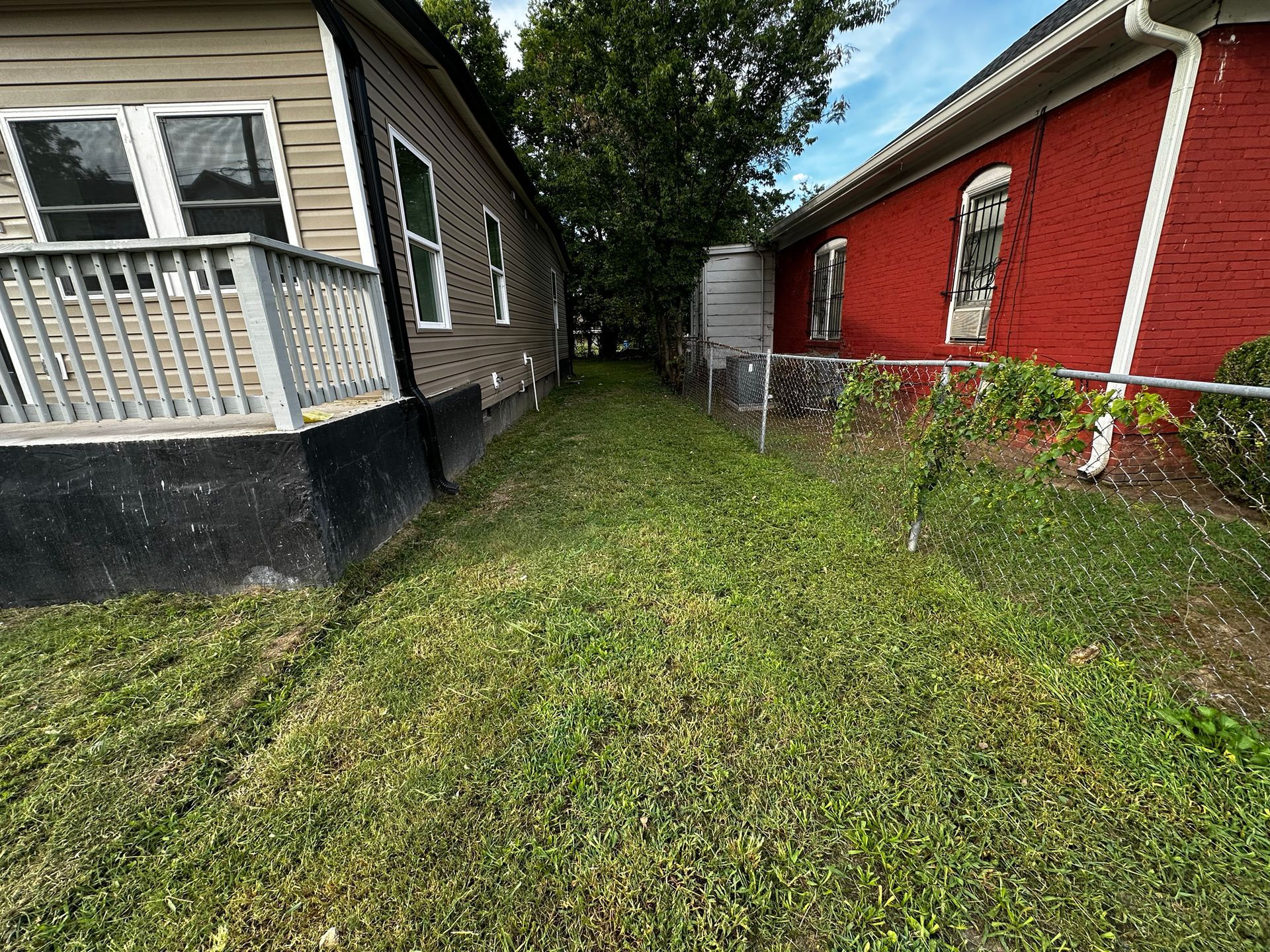 Narrow grassy alley between a tan house with porch and a red brick building. A small shed is visible.