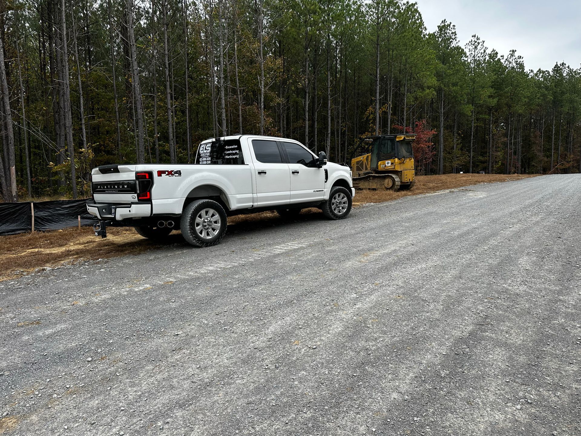 White pickup truck parked on a gravel road near a small bulldozer in a wooded area.