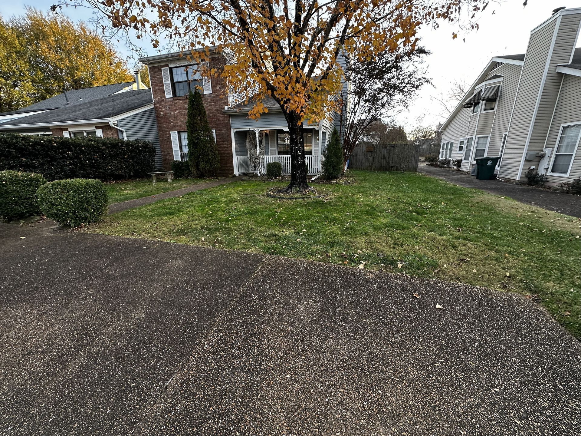 A small house with a porch and yellow tree in front, next to a brick building.