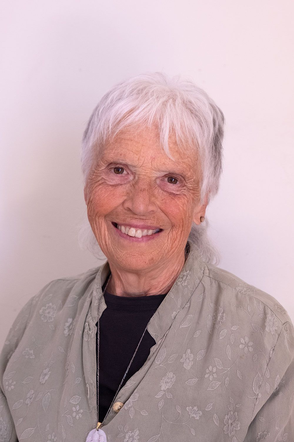 Betsy Rose. Woman with short white hair, hands clasped in front of her face, glasses on. Light green patterned top, indoor setting.