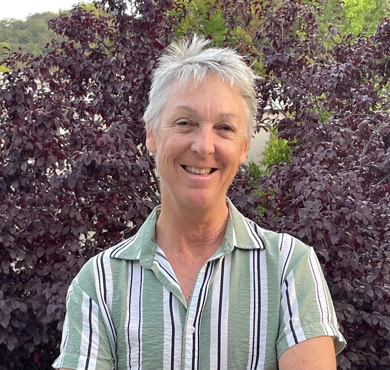 Journey deVries. Woman with short gray hair smiles, wearing green striped shirt, in front of burgundy bush.