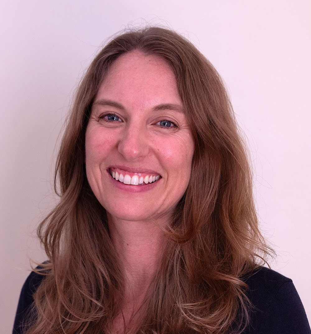 Kate Munding. Woman with blonde hair smiles in front of a tree in a park, with trees and water in the background.