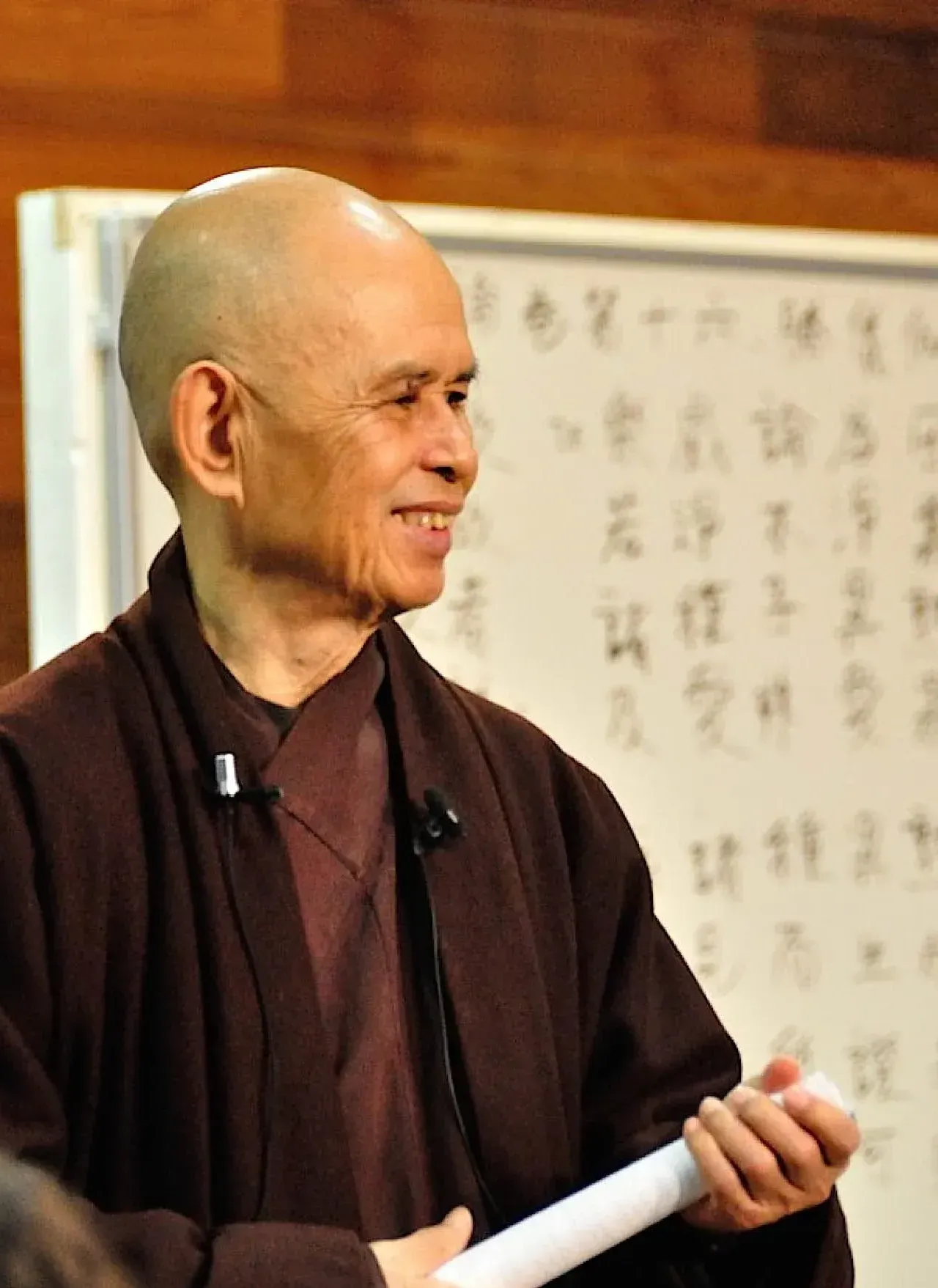 Thich-Nhat-Hanh. A smiling bald monk in brown robes, holding a white scroll, stands by a whiteboard with Asian script.