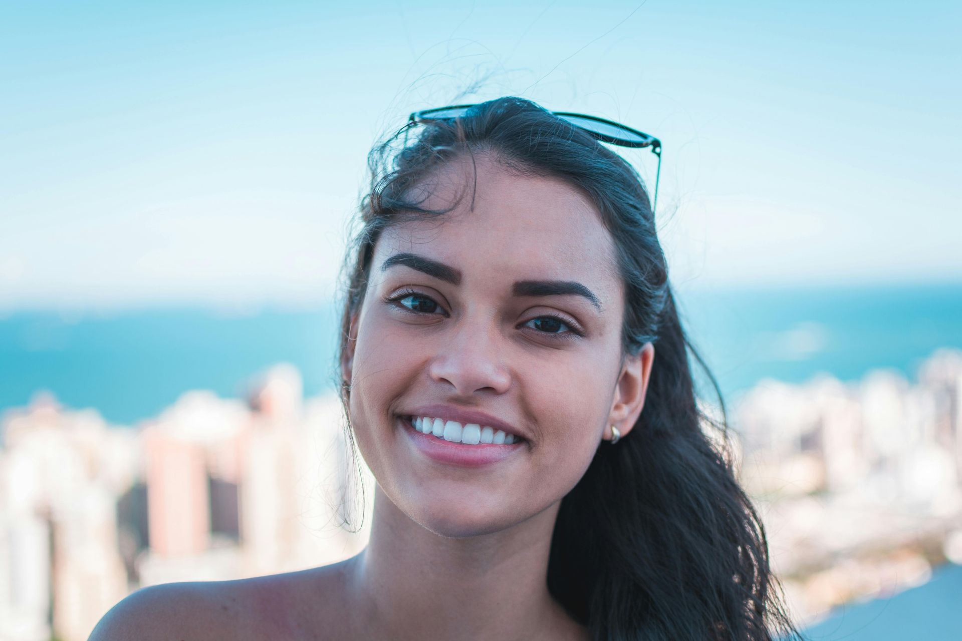 Smiling woman with dark hair and sunglasses atop her head, city and sea in background.