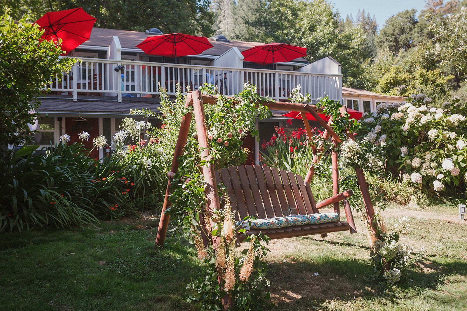 Wooden swing in a garden, red umbrellas on a balcony, surrounded by flowers and greenery.
