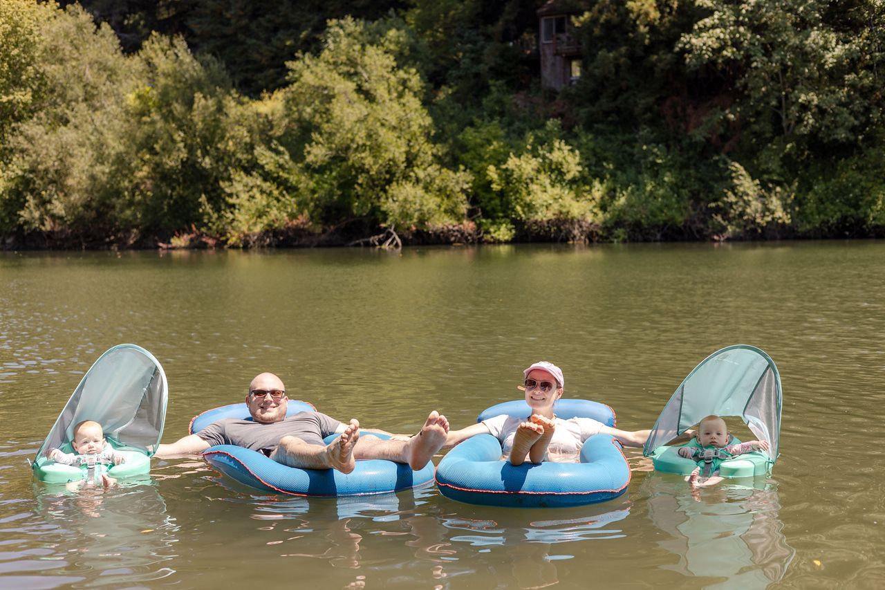 Family floating in a lake on inflatable tubes, with babies in shaded floats. Trees in the background.