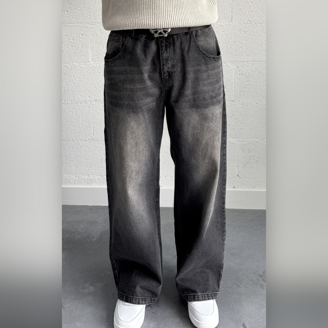 Persona de pie con vaqueros negros extragrandes, suéter blanco y zapatos. Pared gris al fondo.