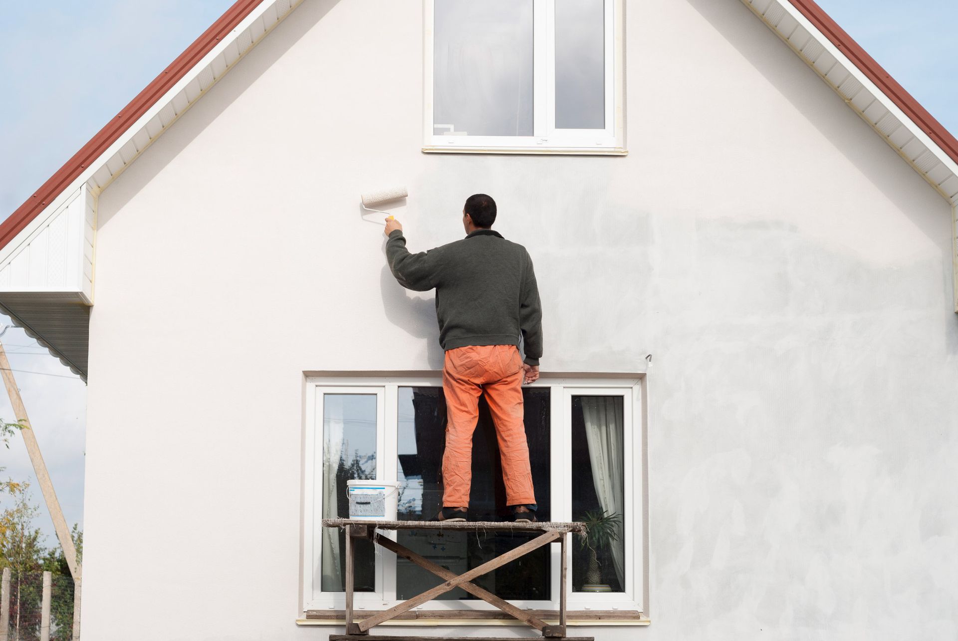 Man paints the exterior of a two-story house. He stands on a scaffold wearing orange pants and a gray sweater.