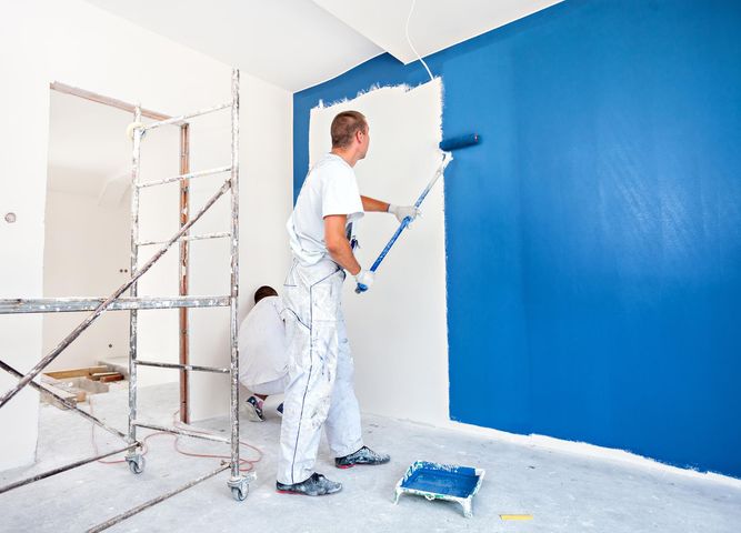 Man painting a blue wall with a roller; another person in the background, room under renovation.
