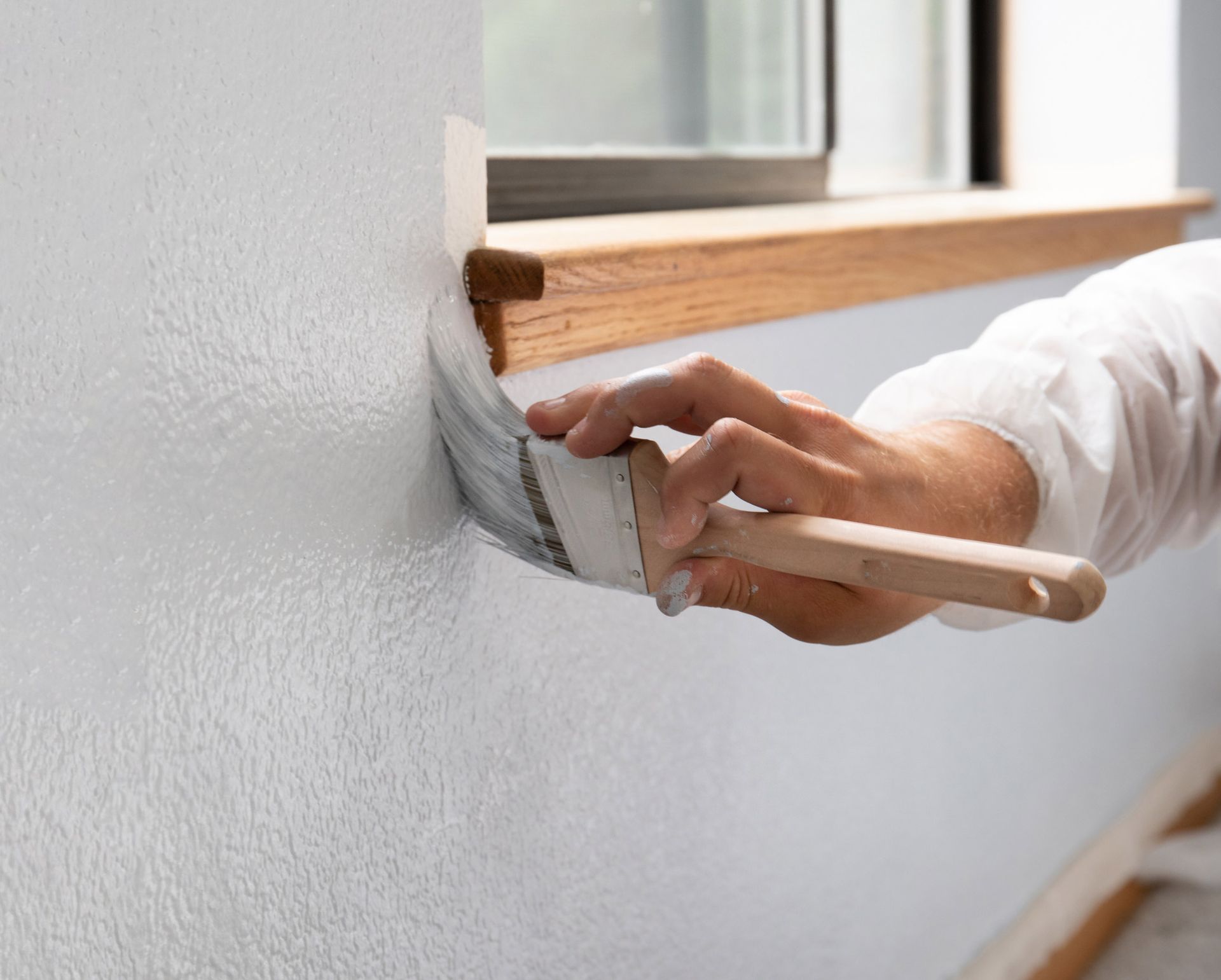 Person painting a wall white with a paintbrush near a window.