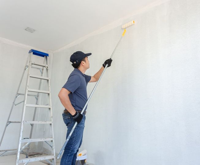 Man painting a white wall with a roller using an extension pole, standing near a ladder.