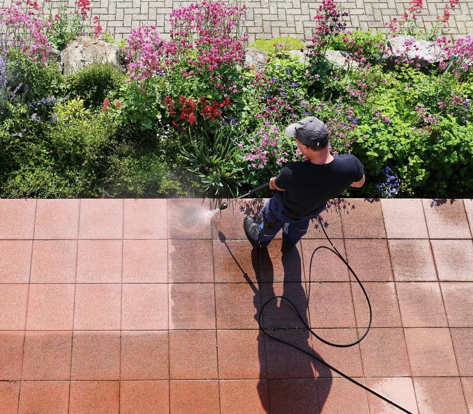 Man power washing a red brick patio next to a lush garden of pink and green plants.