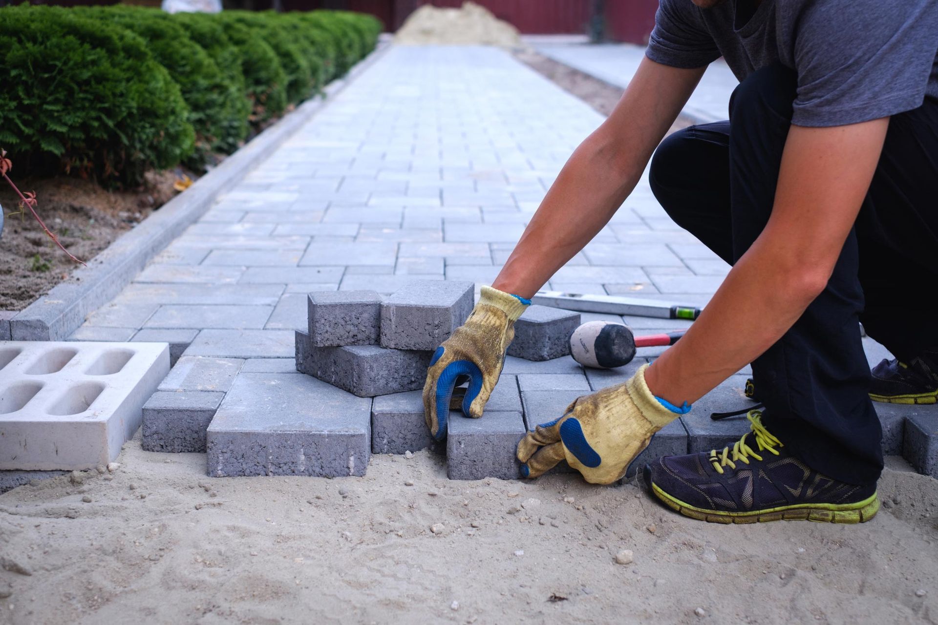 Person laying gray paving stones, kneeling on sand.