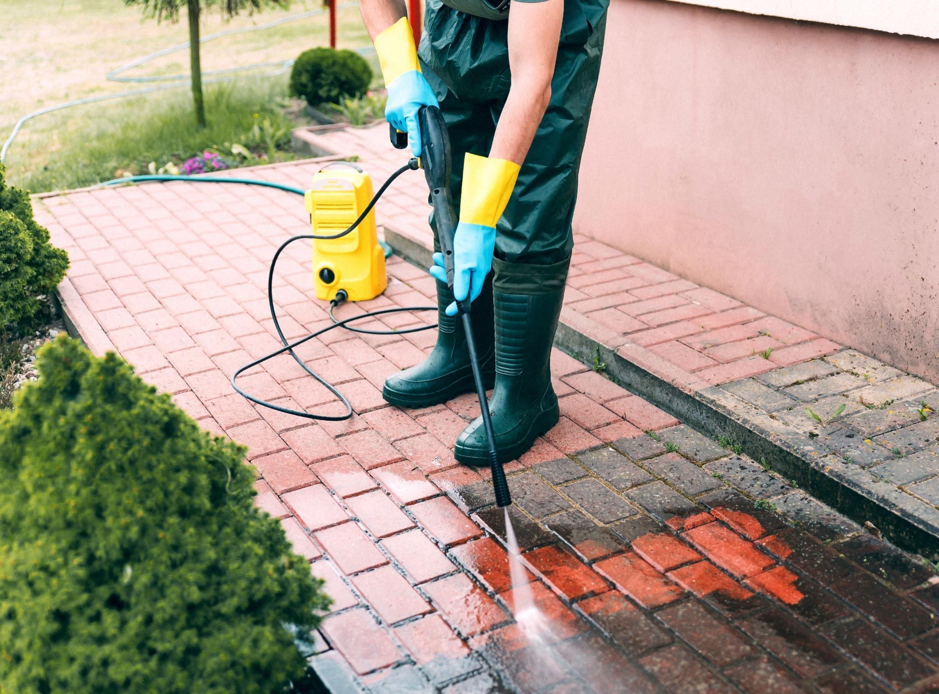 Person power washing a red brick patio with a yellow pressure washer. Wearing green waders and blue gloves.