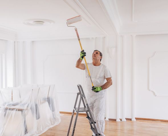 Painter in white coveralls on a ladder, using a paint roller on a ceiling.