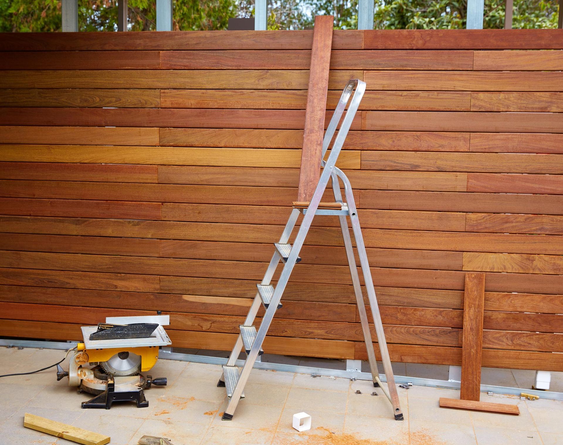 Ladder propped against a wooden fence with lumber and a saw, outdoor setting.